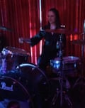 Lara Williams dressed in black playing drums in a darkened room with red curtains behind her