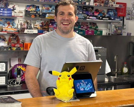 Man stands at counter in shop with games behind and a Pokémon stuffed toy in front