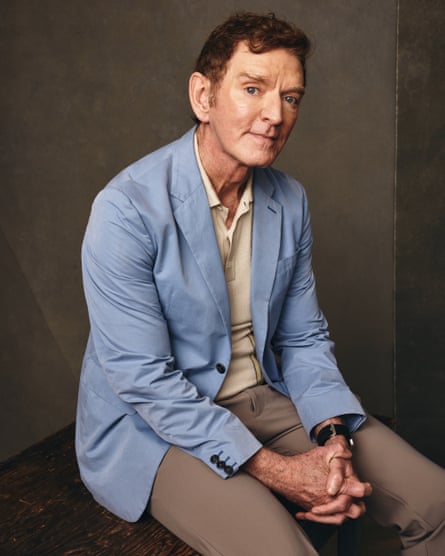 A man wearing a light blue jacket sits with his hands folded on his lap.