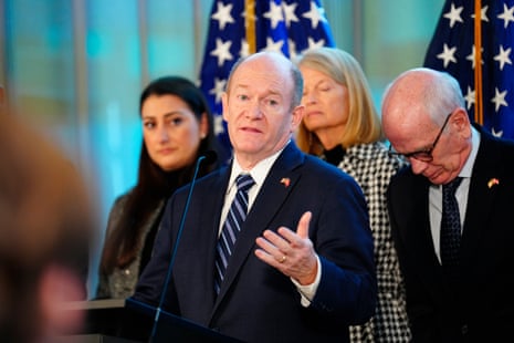 Chris Coons, in a navy blue suit, stands in front of Lisa Murkowski and others before two American flags. Coons is gesturing with his left hand.