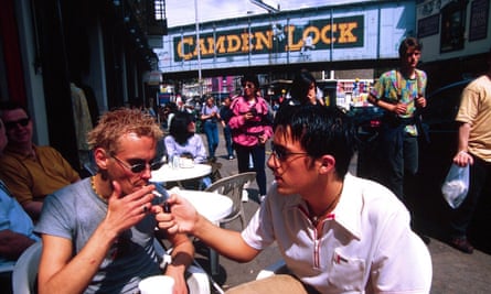 A man lights another man’s cigarette at a cafe table with a bridge behind bearing the name ‘CAMDEN LOCK’ in large letters