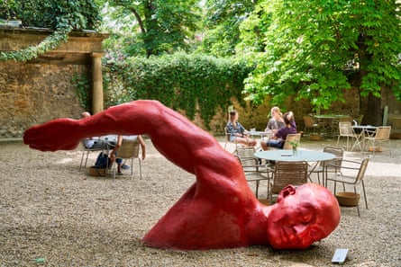 A large red sculpture of a swimmer’s arm, head and torso emerging from the gravel of a courtyard with cafe tables and chairs