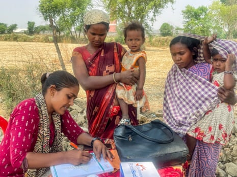 Two Indian women in saris stand with their small children in their arms watching a seated woman makes notes on a pad of paper