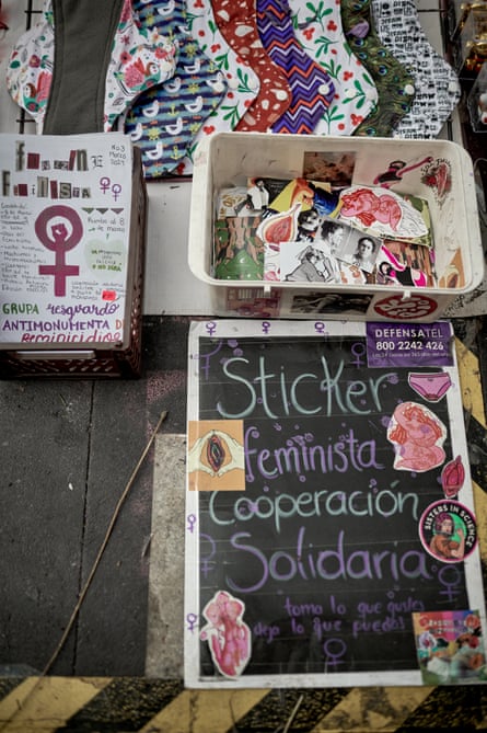 Laid out of the ground are a selection of brightly coloured reusable sanitary towels, a plastic bucket with a jumble of different feminist stickers and a sign showcasing a few of the deigns with a helpline number in the corner.
