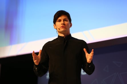 he wears a headset microphone and holds his hands out in a gesticulation. He has short dark hair and is clean-shaven, and wears a black polo-neck jumper.