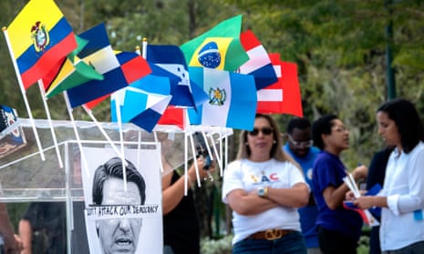 flags from central and south american countries adorn a podium with a picture of ron desantis with the words 'don't attack our democracy'. people stand in the background talking