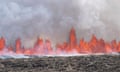 A line of lava launches upwards from bare ground with smoke filling the air above