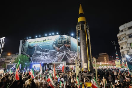 A towering ballistic missile is surrounded by a crowd of people waving Iranian national flags
