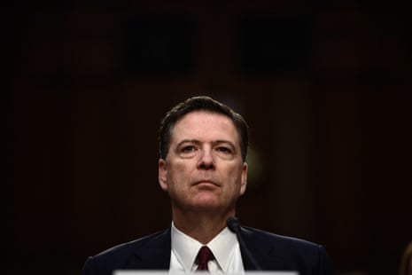 James Comey stands with a serious expression before a microphone at a hearing