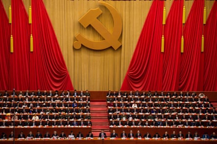 Ranks of officials seated in vast hall with hammer and sickle symbol on the wall