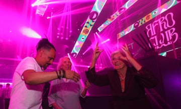 Two older women and one younger man in a nightclub surrounded by pink-purple lighting and colourful graphics. One of the women has both hands in the air.