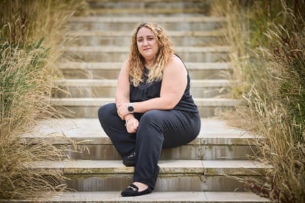 Kirsty-Anne Holmes sits on a flight of steps with tall grass to either side of her. She has long blond curly hair and wears a black sleeveless top and trousers.