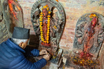 A man with white hair and wearing a black hat places an offering at the foot of a stone statue wearing a flower garland