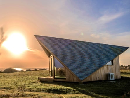 A small wooden cabin with a dramatically shaped roof next to a beach at sunrise