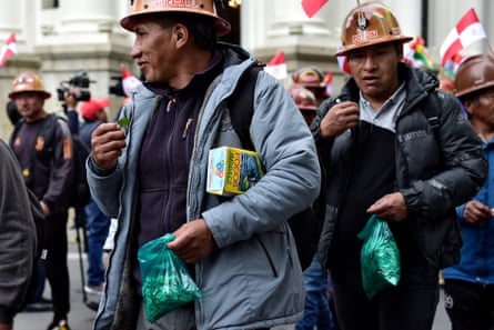 Two Indigenous men in miners’ helmets eat leaves from a bag as they march in a procession