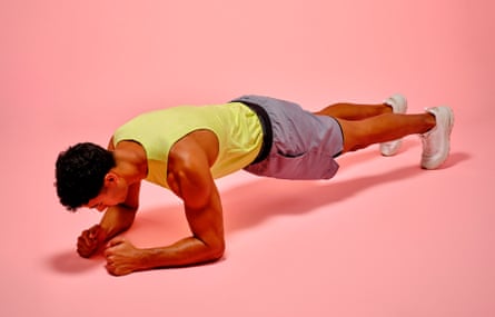 A young man with muscly arms wearing a yellow vest, grey shorts and white trainers, and doing a plank
