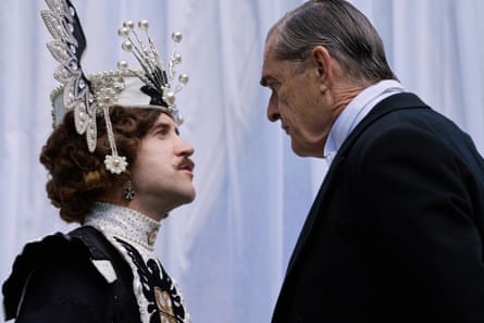Everett, in a black suit, speaks to a moustchioed aristocrat in spangled headpiece