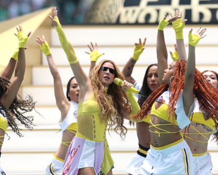Shakira waves her hand in the air along with several back-up dancers, while singing into a mic