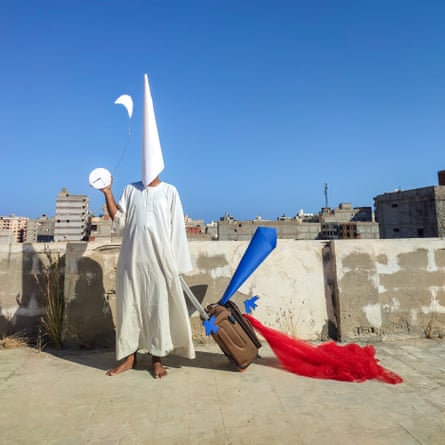 A person in a white conical mask and white robe pulling a suitcase behind them that has a piece of red fabric tied to it