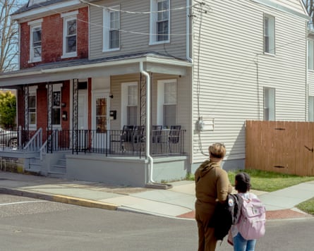 A middle-aged Black woman with a frosted blond bob stands with a maybe 10-year-old Black girl in the middle of an intersection, both looking at a two-story duplex on the corner, that has a shared porch, white siding on the right half, and brick on the left half.