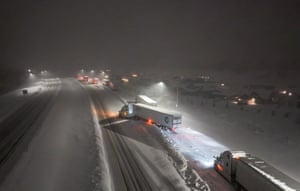 Trucks sit at the side of the snow-covered highway at night. One of them is jutting out into the road