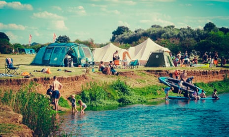 Family tents by a river bank, with kids playing with a dinghy in the river in the sunshine.
