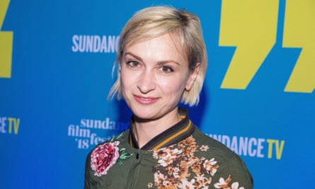 Young white woman with chin-length blong hair and green flowered bomber jacket smiles in front of blue-and-yellow backdrop for a press photo.