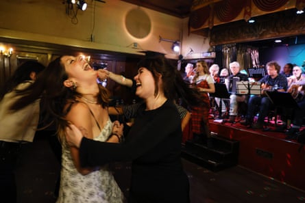 Two young women dancing together and laughing. One, wearing a white dress, has her head held back and mouth wide open in laughter. The folk orchestra plays on the stage.