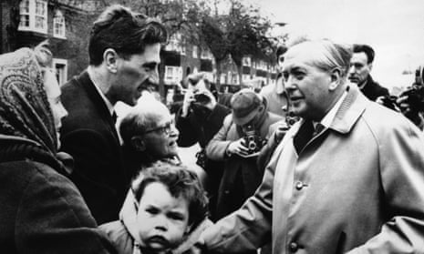 Bland and white photo of Wilson, in a raincoat, talking to a man, woman and child on the street, with a photographer taking a picture in the background