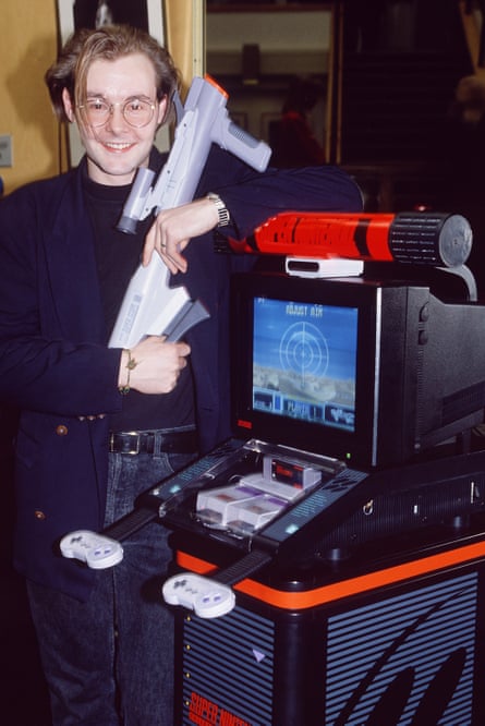 A man with floppy blonde hair and glassed smiles as he holds a toy gun while standing next to an old fashioned video game.