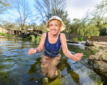 A woman standing waistdeep in water wearing a swimming costume and hat and looking cold.