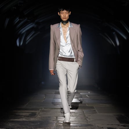 A model wearing a mushroom-brown jacket over a silver open-necked shirt with light grey trousers