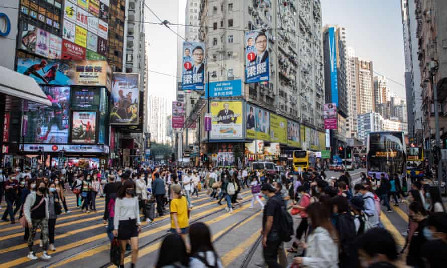 Crowds of people cross street. Tall buildings display election posters.