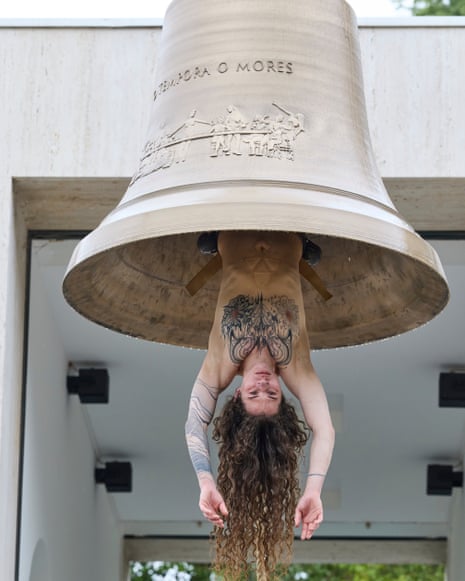 A person performs an upside-down handstand beneath a large bell, with long curly hair hanging down.