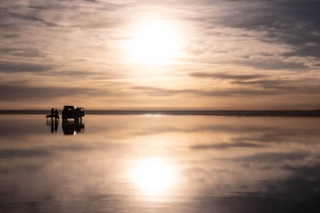 A hazy sun over a vehicle and several people standing on salt flats covered with a thin layer of water