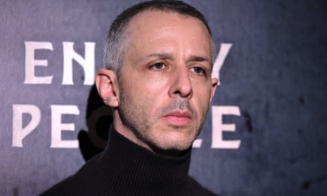 Middle aged white man with trip black hair gray on sides, wearing black turtleneck, looks serious in front of press background.