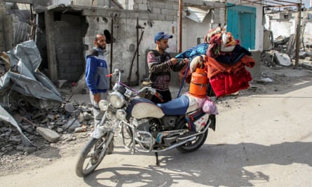 A Palestinian man loads his belongings on a motorcycle as another man looks on; clothes, blankets and a gas cylinder are strapped to the back of the motorbike. Both men are quite young and wear zipped-up sporty sweatshirts; they are in a street in front of a line of ruined, bombed-out houses with piles of rubble around them.