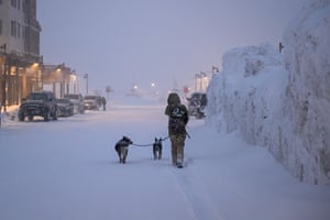 A woman walks away from the camera down a snow-covered street, with dogs beside her