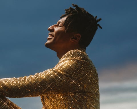 Jon Batiste in a gold sequined outfit sits on concrete looking up at a sunset sky