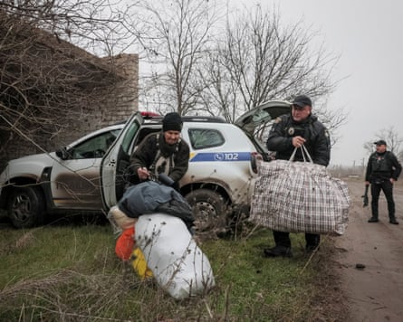 A police officer and a civilian grapple with large bags near an official vehicle with the hatch open, while another officer dangling a rifle waits watching the surroundings