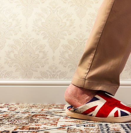 A brown-trousered leg against a wallpapered wall and skirting board, stepping out of frame, with the bare foot wearing a union jack slipper