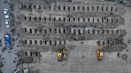 Aerial shot of five rows of graves, about 20 in a row, being dug by bucket-loader tractors, with those to be dug marked out in white lines on the grey ground.
