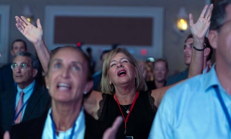 In a large conference hall, we see a blonde white middle-aged woman, in full makeup, a black dress, and a red lanyard around her neck, lift her hands, eyes closed, appearing to sing, with others around her also appearing to joyfully sing.