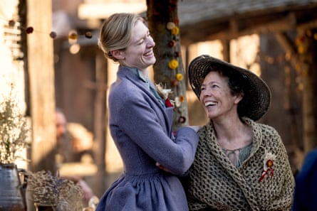 A tall blond woman in a blue dress and a shorter brunette woman wearing a coat and hat. Both smiling at each other