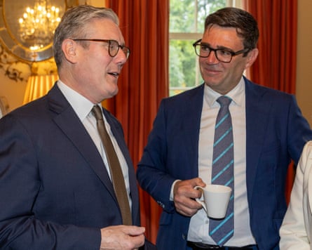 Burnham, holding a cup, listens to Starmer speaking.