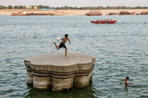 A boy on top of a plateau plays with a football on the banks of the river Ganges at Scindia Ghat on a hot summer’s day