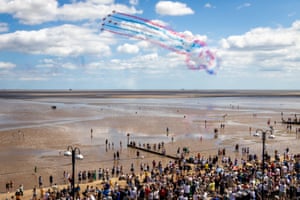 Seven aircraft fly over a beach, leaving red, white and blue smoke trails