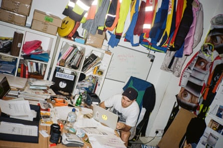 Johnny Banger, con indosso un berretto da baseball e una maglietta, guarda un laptop con un adesivo con una faccia felice nel suo studio, dove ci sono molti libri, scatole, tazze, documenti e grucce.