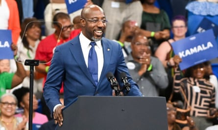 Black man with bald head and blue suit smiles and holds lectern.