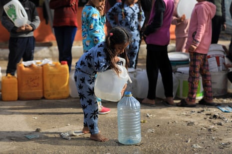 A small girl with bare feet carefully pours water from one container to another as other children queue with jerrycans